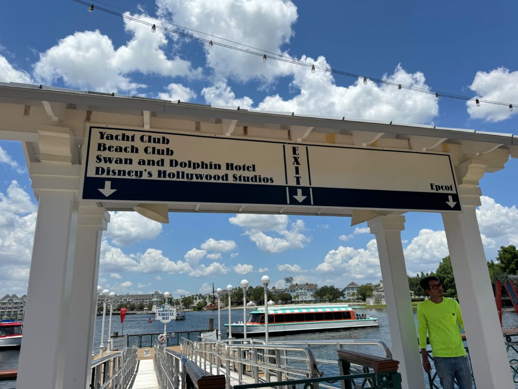 Sign at a boat dock displaying directions to yacht club, beach club, swan and dolphin hotel, and disney's hollywood studios under a sunny sky.